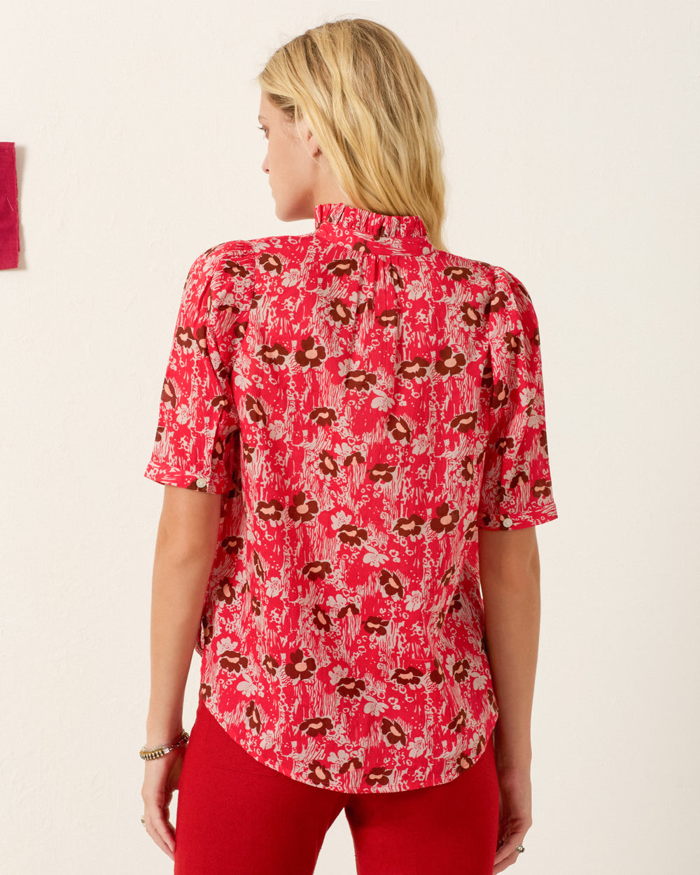 A woman with blonde hair wears the Winnie Rougewood Shirt, a red floral puff-shoulder blouse, paired with red pants and stands with her back to the camera against a light background.