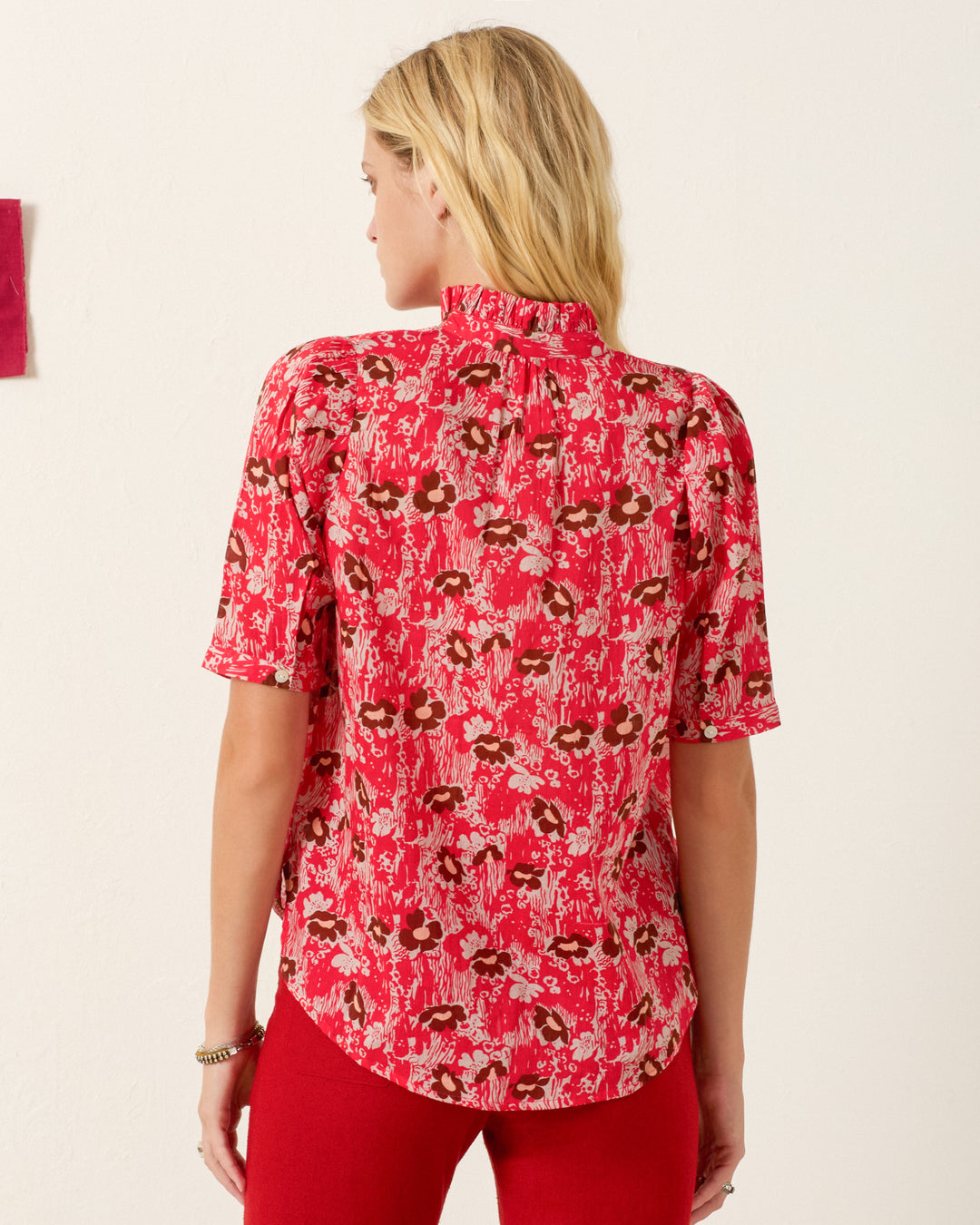 A woman with blonde hair wears the Winnie Rougewood Shirt, a red floral puff-shoulder blouse, paired with red pants and stands with her back to the camera against a light background.