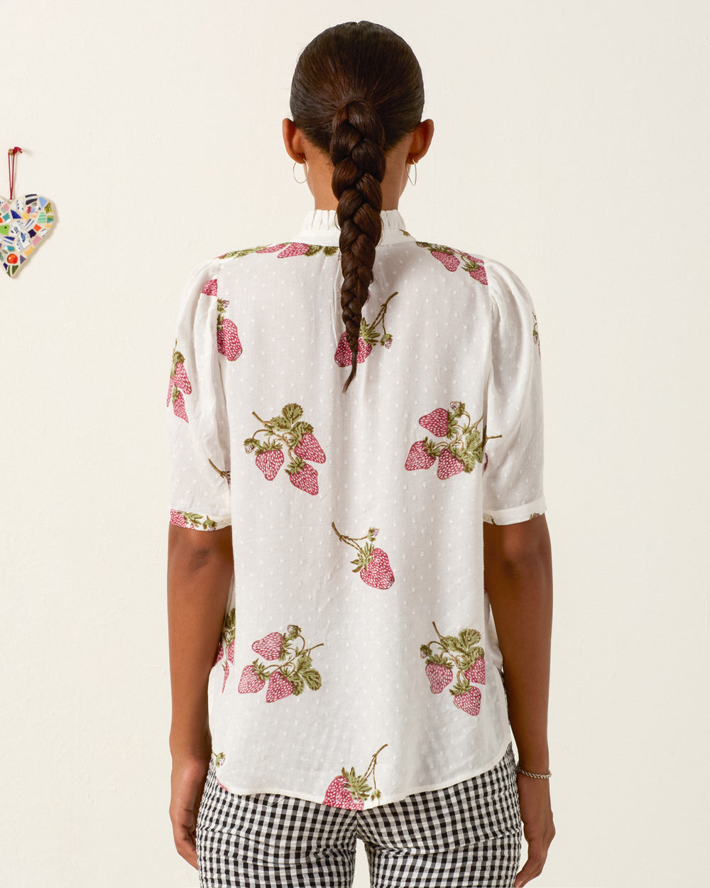 A person with a long braid stands facing away, wearing the Winnie Strawberry Dot Shirt and black-and-white checkered pants. A small heart decoration hangs on the wall to the left.