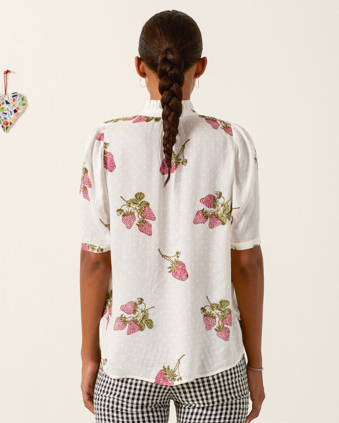 A person with a long braid stands facing away, wearing the Winnie Strawberry Dot Shirt and black-and-white checkered pants. A small heart decoration hangs on the wall to the left.