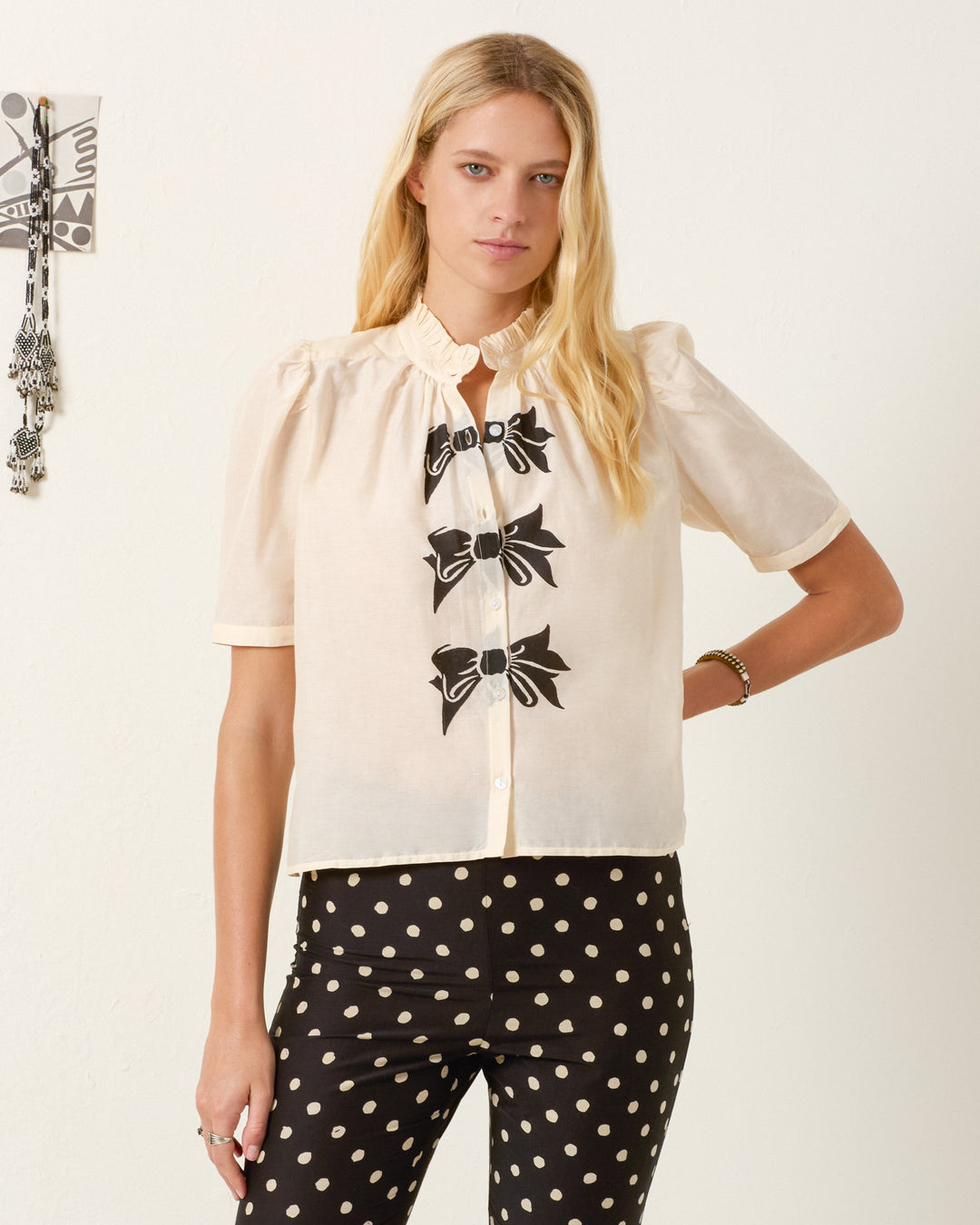 A woman wearing the Winn Black Tie Shirt, a short-sleeve cream blouse with trompe l'oeil bows, and black polka dot pants stands against a plain background.