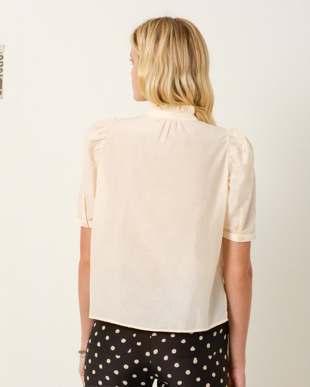 A woman with long blonde hair is shown from the back, wearing the light beige Winn Black Tie Shirt adorned with trompe l'oeil bows and black pants with white polka dots.