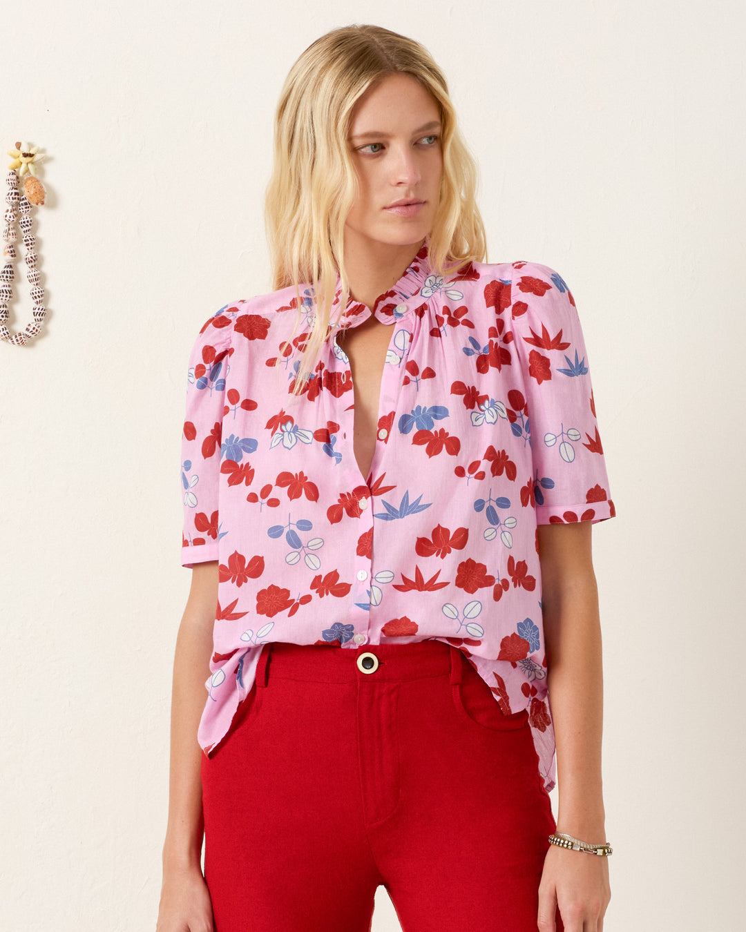 A woman wears the Winnie Tutu Bloom Shirt—a pink botanical cotton voile top with a ruffled collar—paired with high-waisted red pants, standing against a plain light wall.