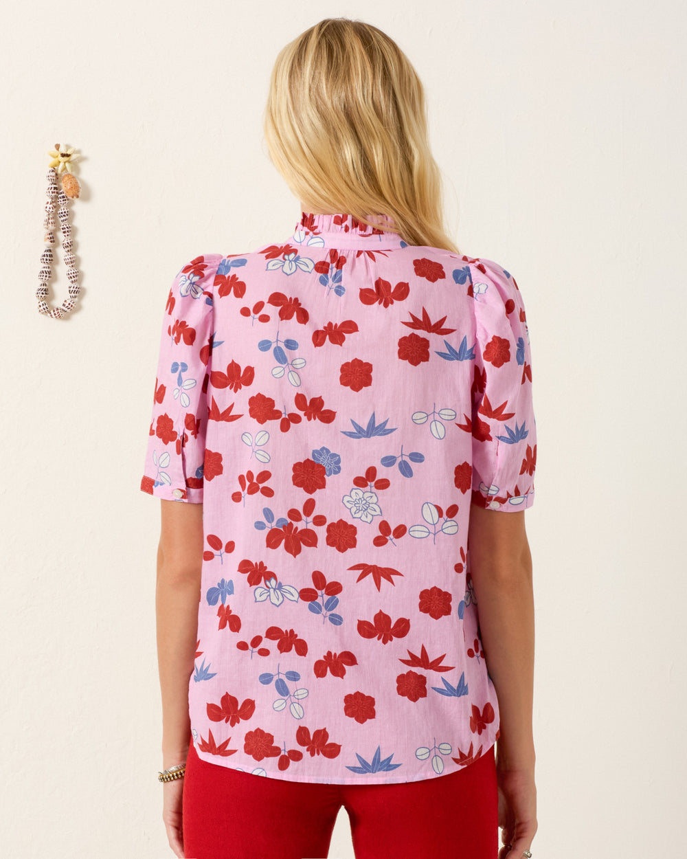 A woman with long blonde hair faces away, wearing the Winnie Tutu Bloom Shirt—a pink cotton voile top with red and blue botanical motifs—paired with red pants, standing against a light-colored wall.