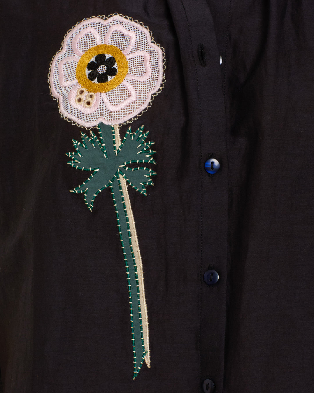 The Winn Pressed Daisy Shirt is a black cotton-silk blend with blue buttons, featuring an embroidered handmade daisy—green leaves and stem, pink, white, and yellow flower head—on the left side.