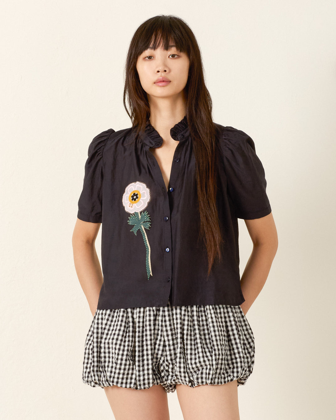 A woman stands against a plain wall, wearing the Winn Pressed Daisy Shirt and black-and-white checkered shorts.
