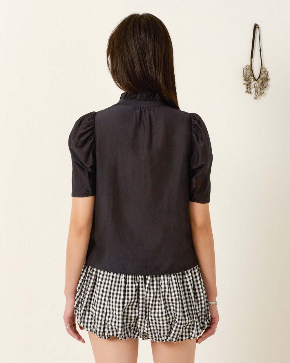 A woman faces away from the camera, wearing the Winn Pressed Daisy Shirt and a black-and-white checkered skirt, with a wall decoration on the right.