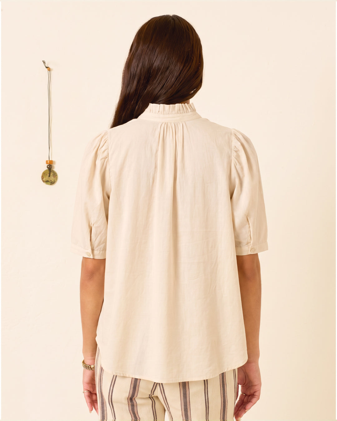 A woman stands facing away, wearing the Winnie Stone Shirt—a light beige cotton voile top featuring short puff shoulders and a ruffled collar—paired with striped pants. A small hanging decoration is on the wall to her left.