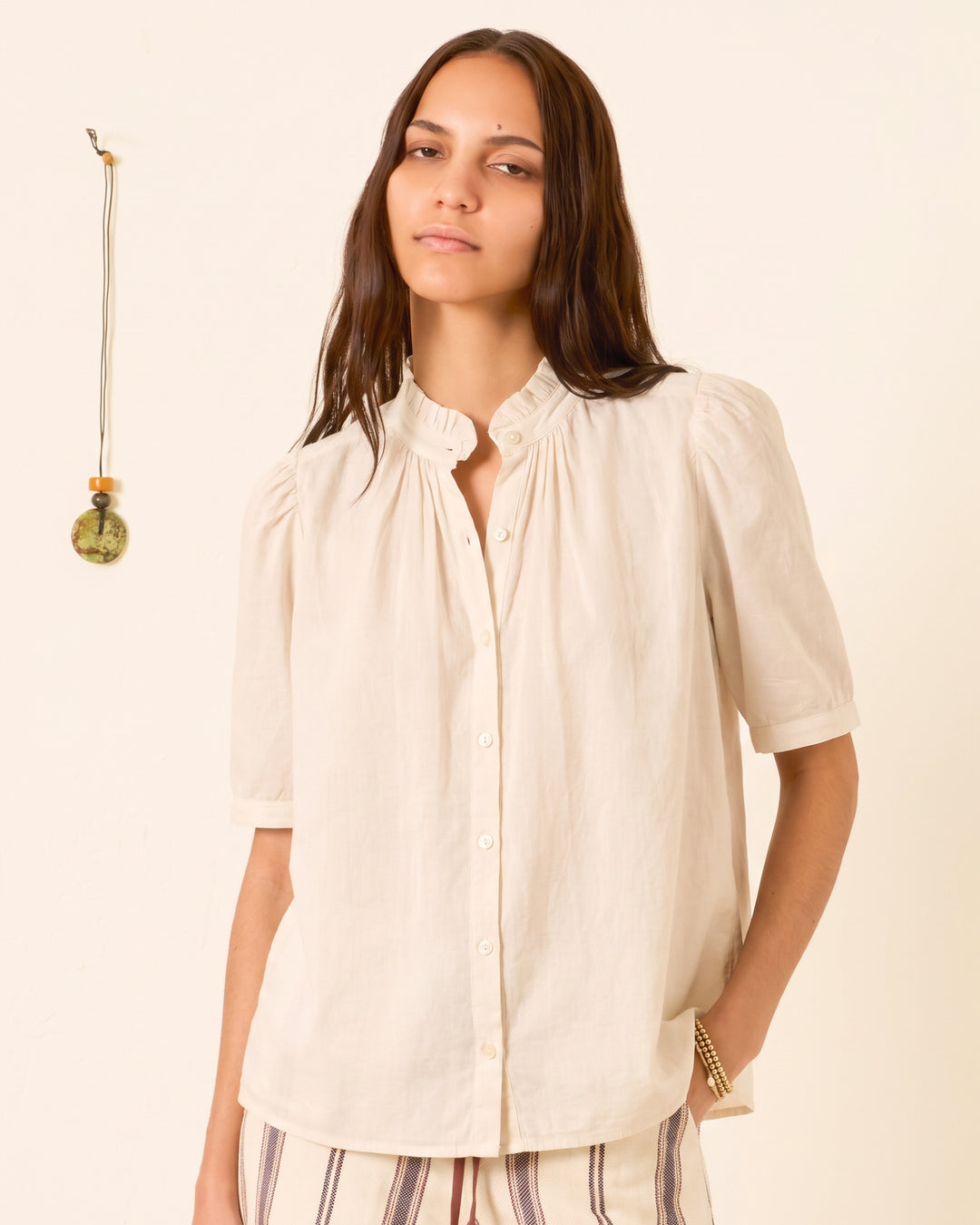 A person is wearing the Winnie Stone Shirt—a cream-colored cotton voile shirt with short sleeves and puff shoulders—standing against a light background with a hanging decorative object on the wall.