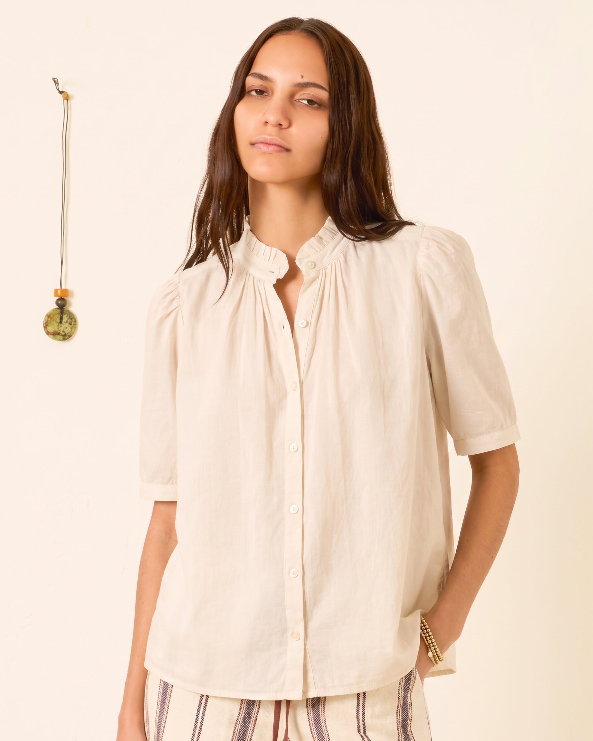 A person is wearing the Winnie Stone Shirt—a cream-colored cotton voile shirt with short sleeves and puff shoulders—standing against a light background with a hanging decorative object on the wall.