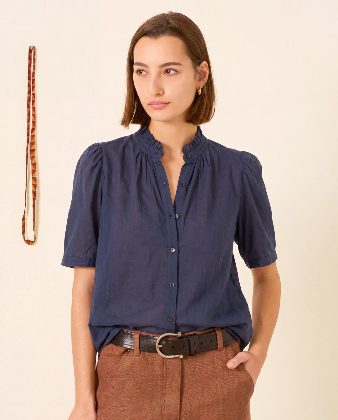 A woman with straight brown hair, dressed in the Winnie Deep Sea Shirt and brown pants, stands against a plain background. A patterned lanyard hangs on the wall.