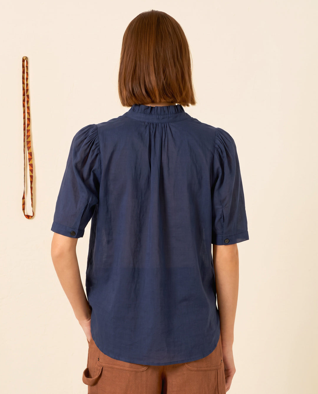 A person with short brown hair, wearing the Winnie Deep Sea Shirt—a navy cotton voile top with a ruffled collar—and brown pants stands facing away from the camera; a patterned lanyard hangs on the wall nearby.