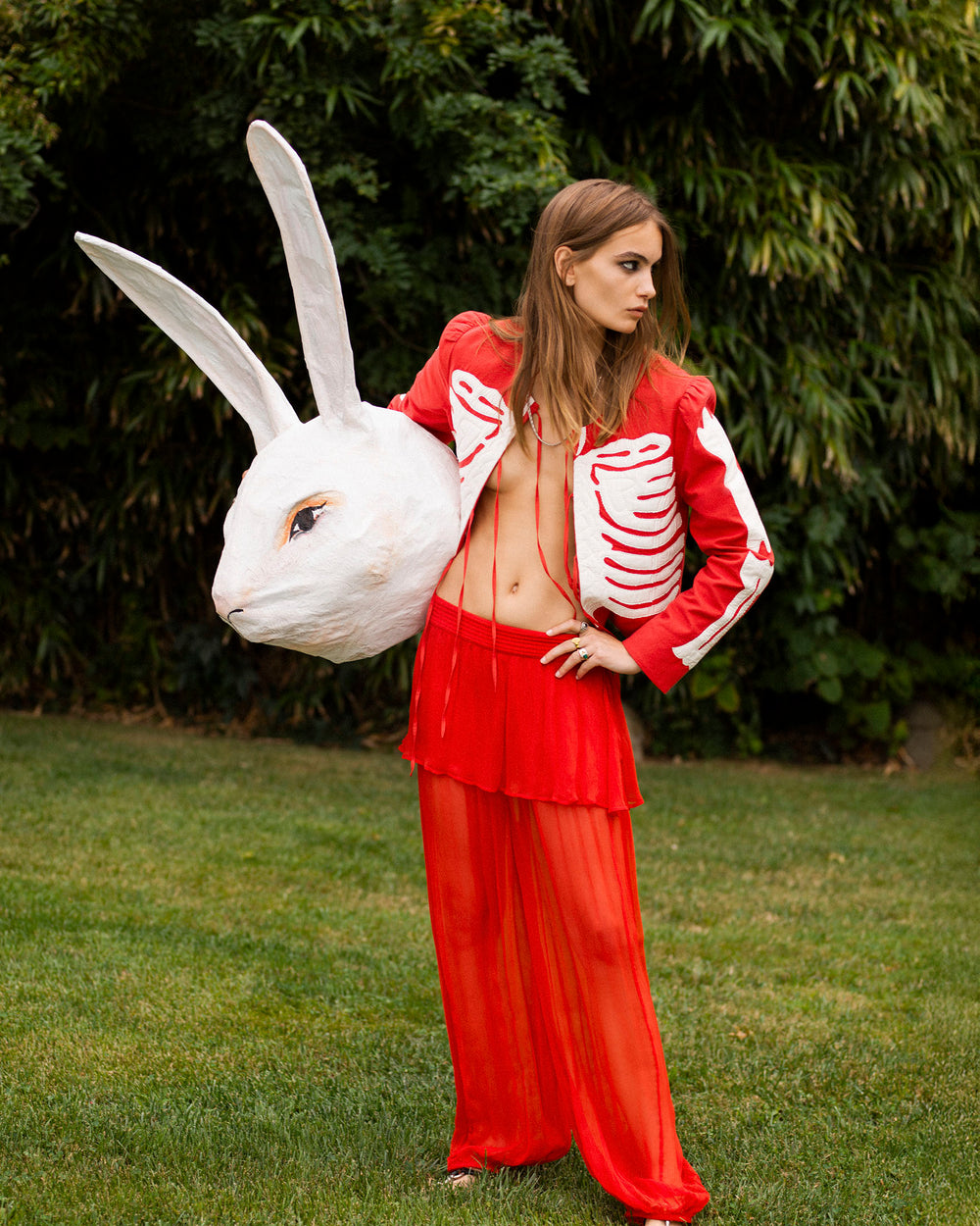 A person wearing the Dorian Ibis Bone Jacket and sheer red pants stands on grass, holding a large white rabbit head prop. Dense green foliage forms the background.