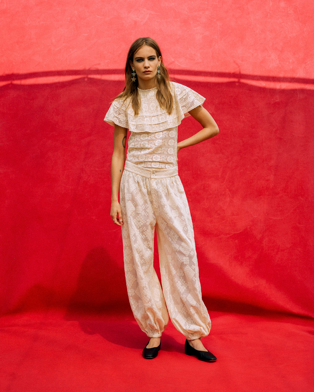 A woman stands against a red background wearing the Margot Ecru Tetris Lace Pant and a matching white ruffled geometric lace top, paired with black shoes.