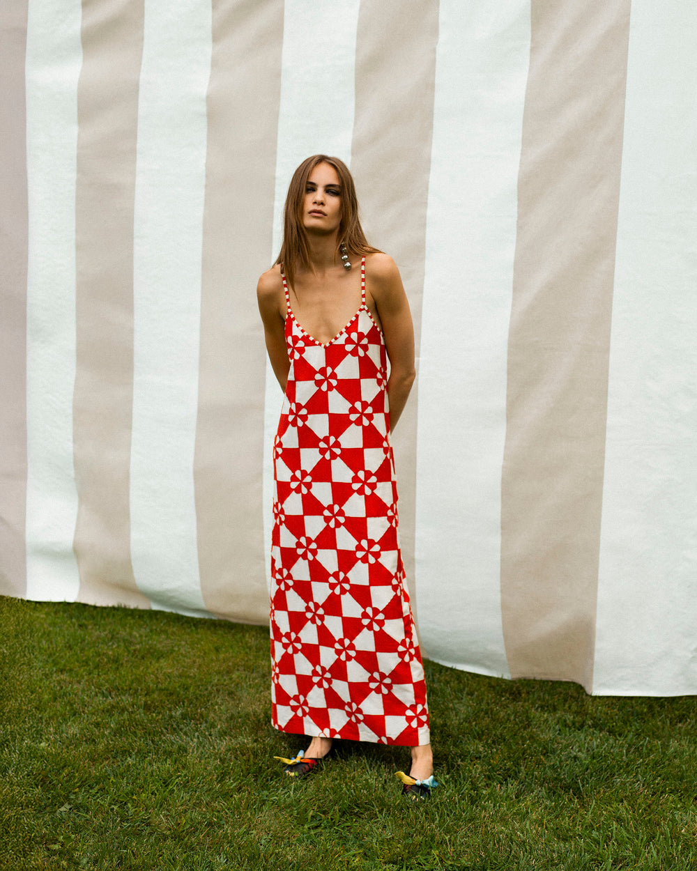 A woman stands on grass before a striped backdrop, wearing the Spice Ibis Jubilee Dress—a sleeveless red and white geometric patchwork slip dress—paired with flat shoes.
