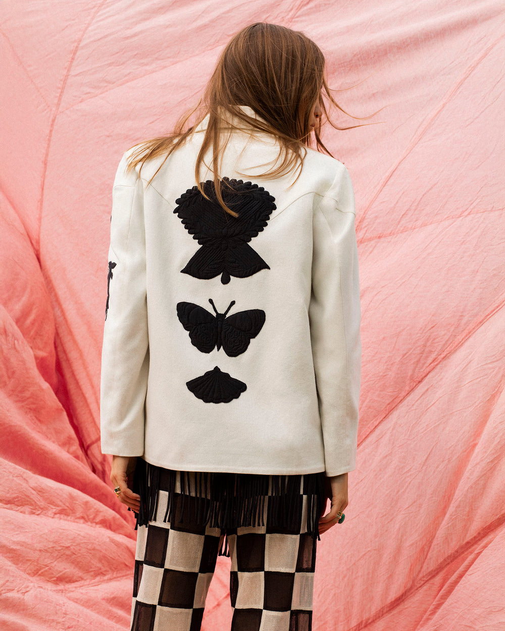 A person stands with their back to the camera, wearing the Jude Pearl Reverie Jacket featuring black butterfly appliqué motifs and checkered pants, in front of a pink fabric backdrop.
