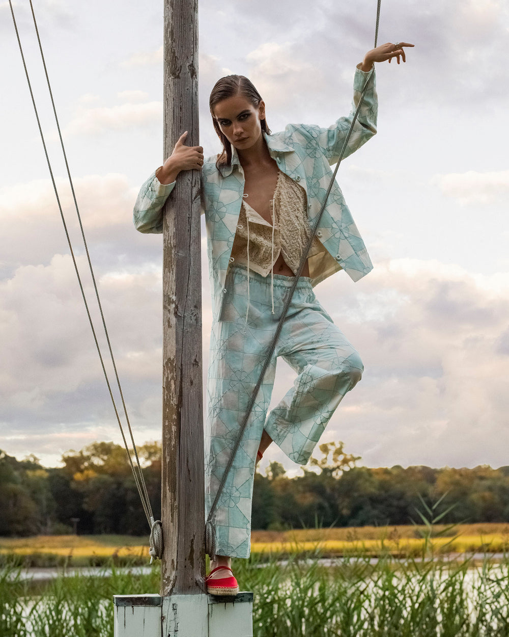 A woman wearing the Jude Sky Jubilee Jacket poses outdoors by a wooden pole near water, with cloudy skies and greenery in the background.