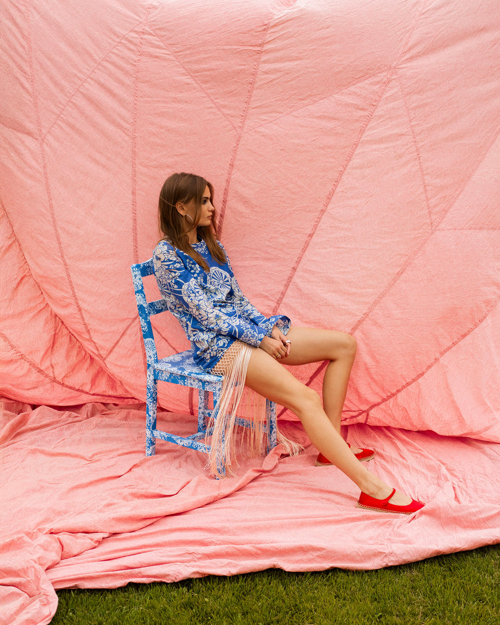A woman in the Narene Royal Spoke Dress and red shoes sits sideways on a blue chair against a large pink fabric backdrop on grass.