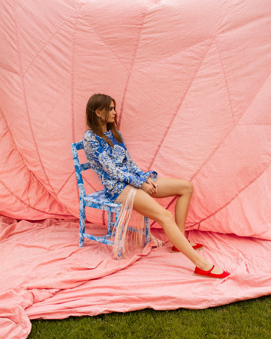 A woman in the Narene Royal Spoke Dress and red shoes sits sideways on a blue chair against a large pink fabric backdrop on grass.