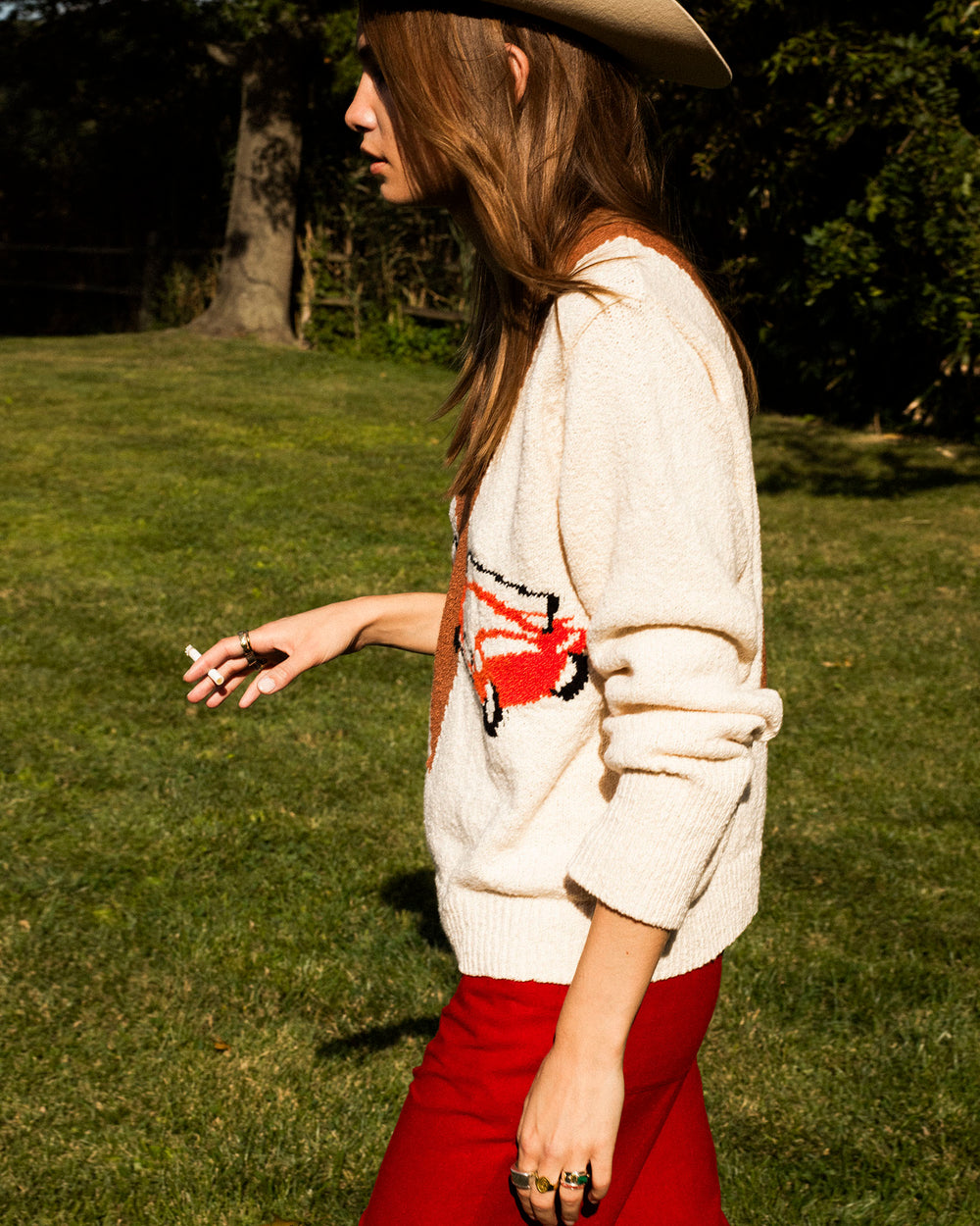 Wearing the Axel Vintage Cruiser Sweater, red pants, and a cowboy hat, a person walks on grass holding a cigarette.