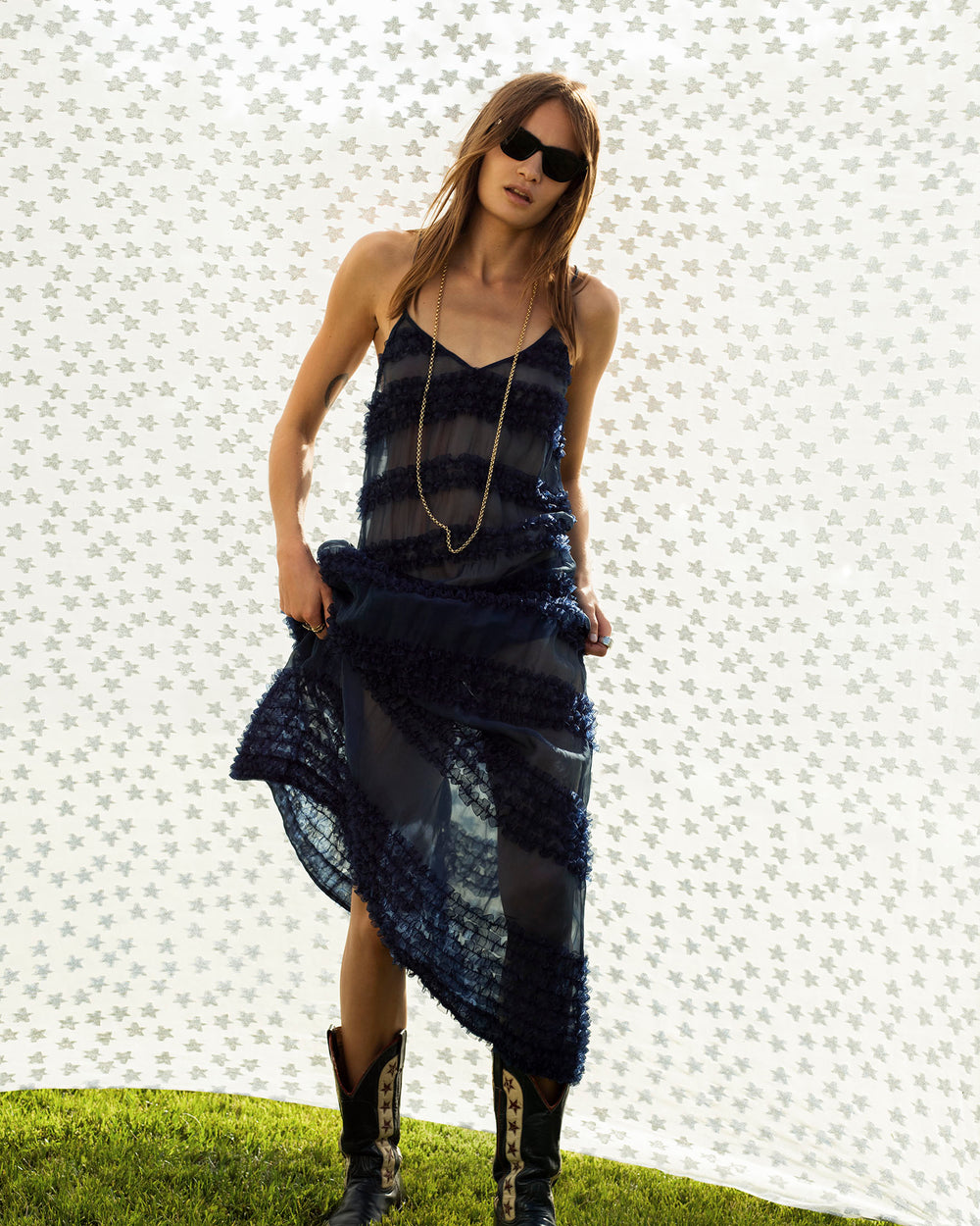 A woman stands outdoors in front of a patterned white backdrop wearing the Spice Market Jester Eve Dress, styled with a long necklace, sunglasses, and boots.