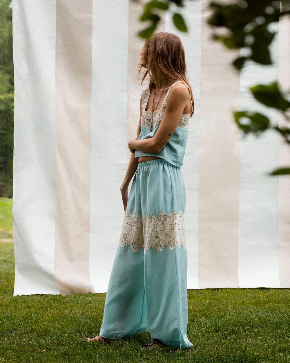 A person stands on grass facing sideways in front of a striped fabric backdrop, wearing Margie Sky Prima Lace Pant.