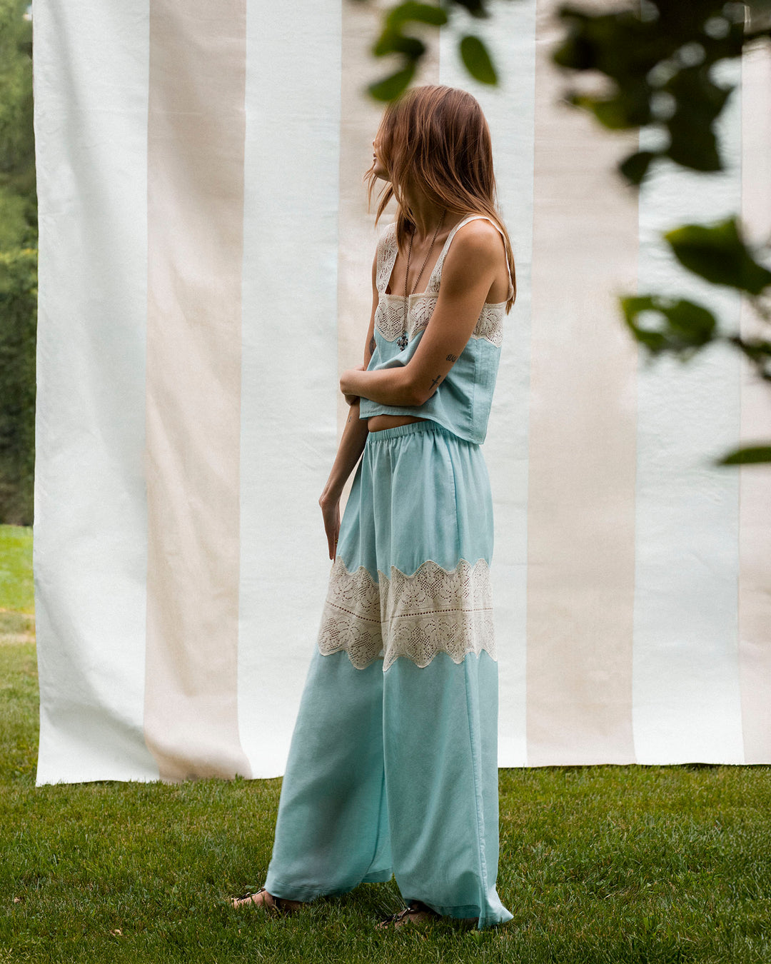 A person stands on grass facing sideways in front of a striped fabric backdrop, wearing Margie Sky Prima Lace Pant.