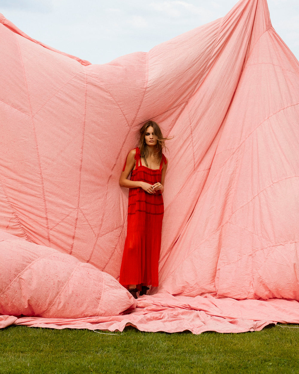 A woman wearing the Emrys Ibis Chiffon Dress with tie shoulder straps stands outdoors on green grass in front of draped pink fabric.
