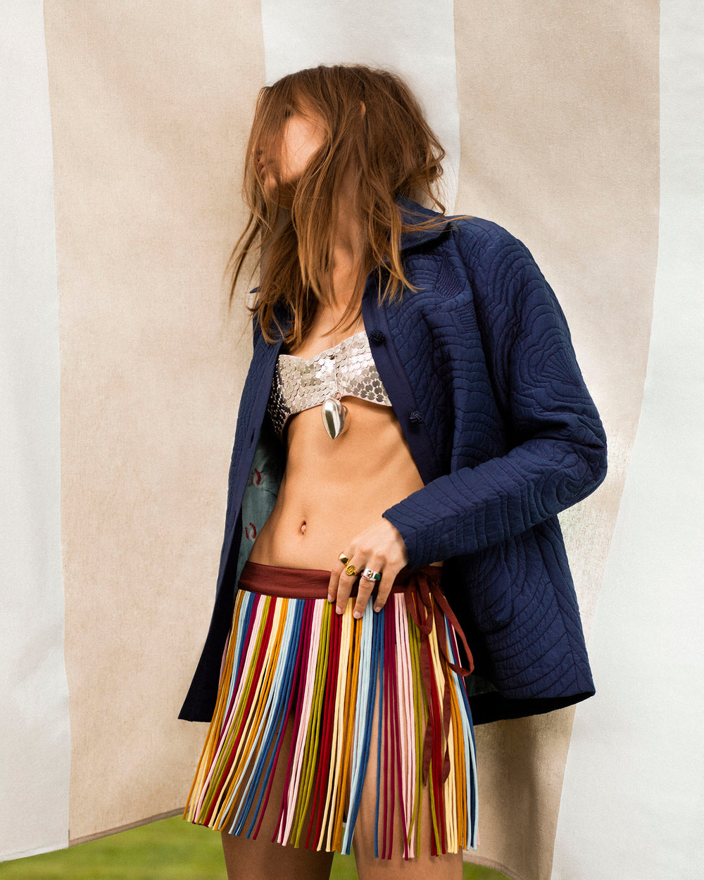 Wearing a dark blue jacket, metallic bralette, and the Tessa Candy Fringe Skirt, a person stands before a striped fabric backdrop, channeling the playful spirit of vintage circus style.