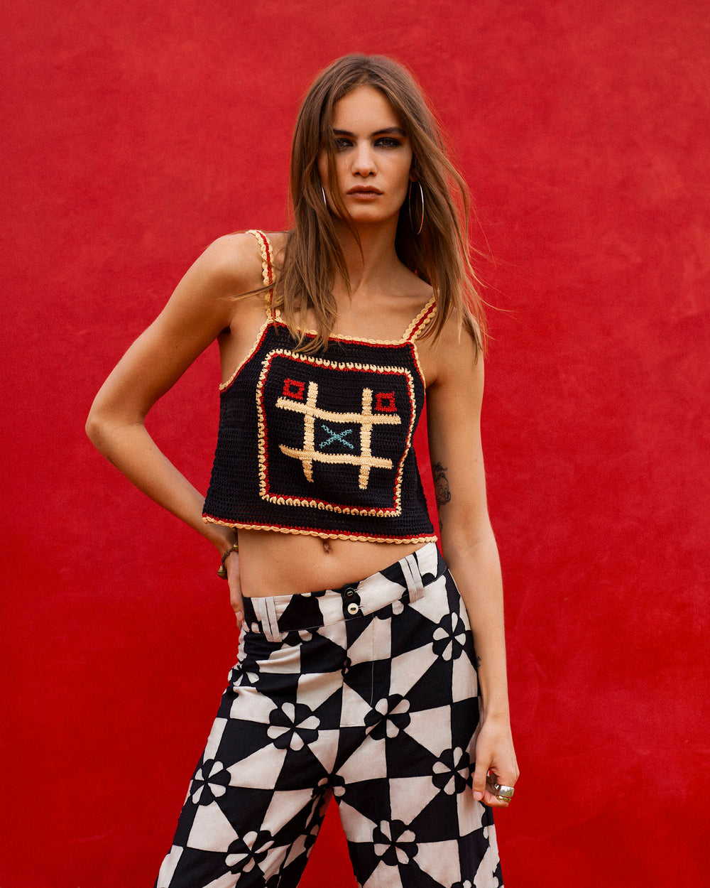 A person stands against a red background, wearing the artisan-made Elide Tic Tac Toe Top—a black hand-crocheted cotton top with a geometric pattern—and black-and-white floral patterned pants.