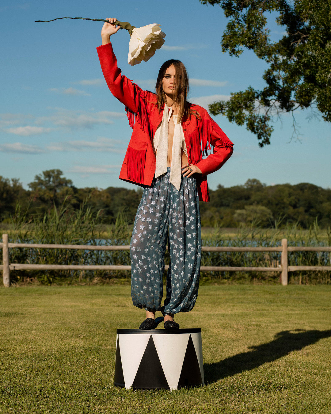 A woman in a Western-inspired red Jude Ibis Fringe Jacket and star-patterned pants stands on a circus pedestal outdoors, raising a large white artificial flower overhead.