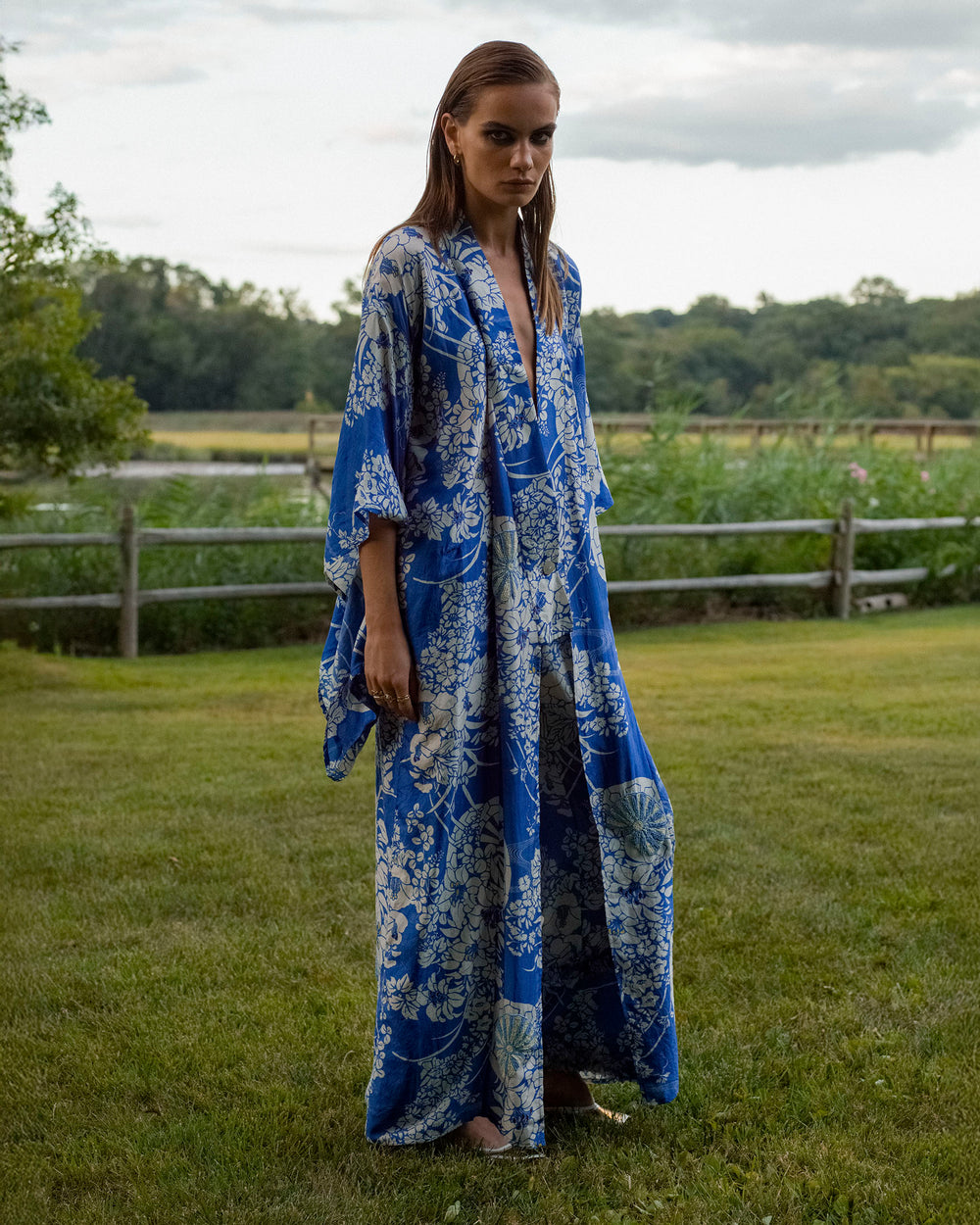 A person stands on a grassy lawn wearing the Grace Royal Spoke Kimono Dress in blue and white silk, with trees and a wooden fence in the background.