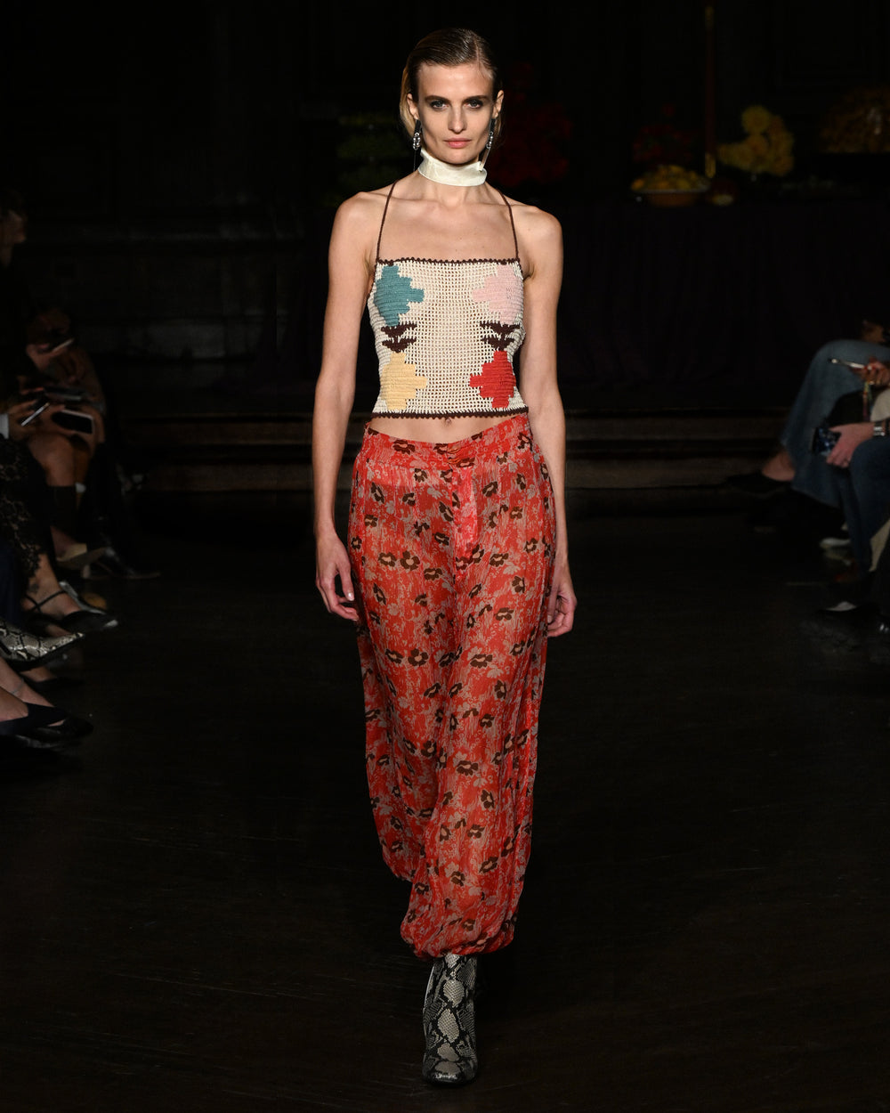 A model walks the runway in the Eliza Daisy Chain Top, a hand-crocheted cotton piece with a geometric pattern, styled with loose red floral pants and snakeskin boots.