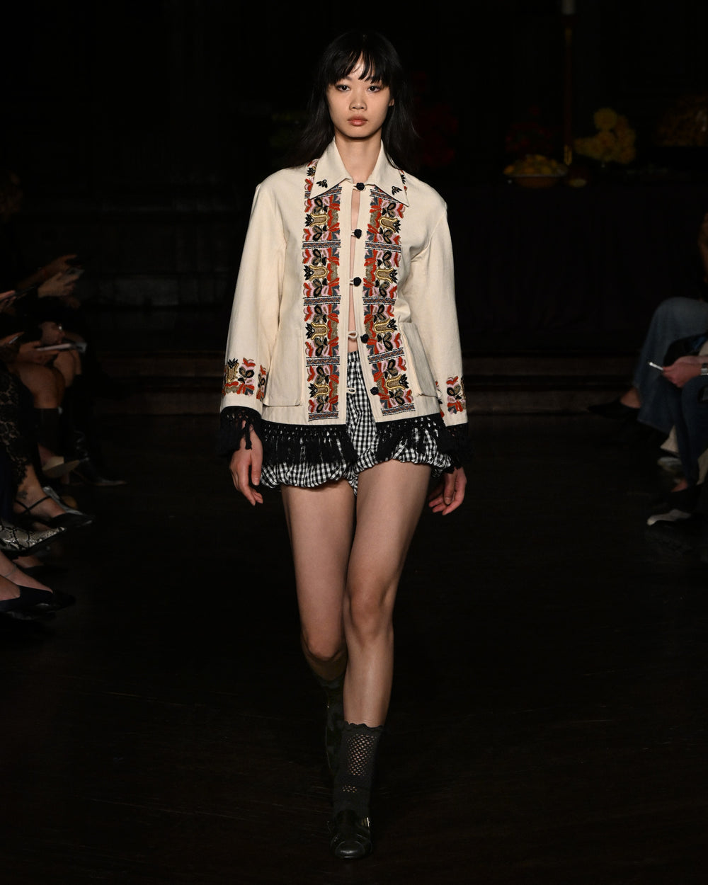 A model walks the runway in the Briar Sideshow Check Short, paired with a cream jacket featuring red and black embroidery, and black boots.