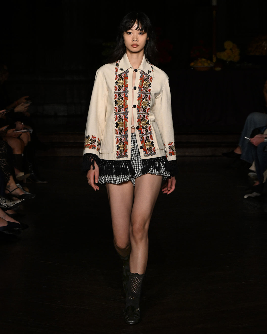 A model walks the runway in the Briar Sideshow Check Short, paired with a cream jacket featuring red and black embroidery, and black boots.