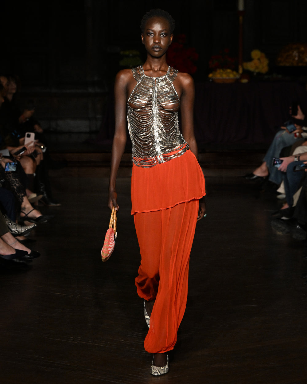 A model walks the runway in a metallic chainmail-style top and Evangeline Ibis Chiffon Pant, holding a small bag, with an audience seated on both sides.