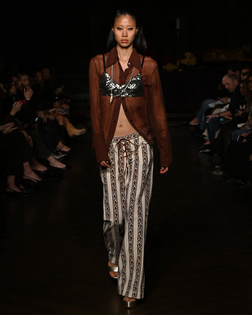 A model walks the runway in a sheer brown shirt, a sequined bra top, and the Cooper Climbing Vine Pant—hand-block-printed khadi cotton pants with a flared leg—as the audience watches from both sides.