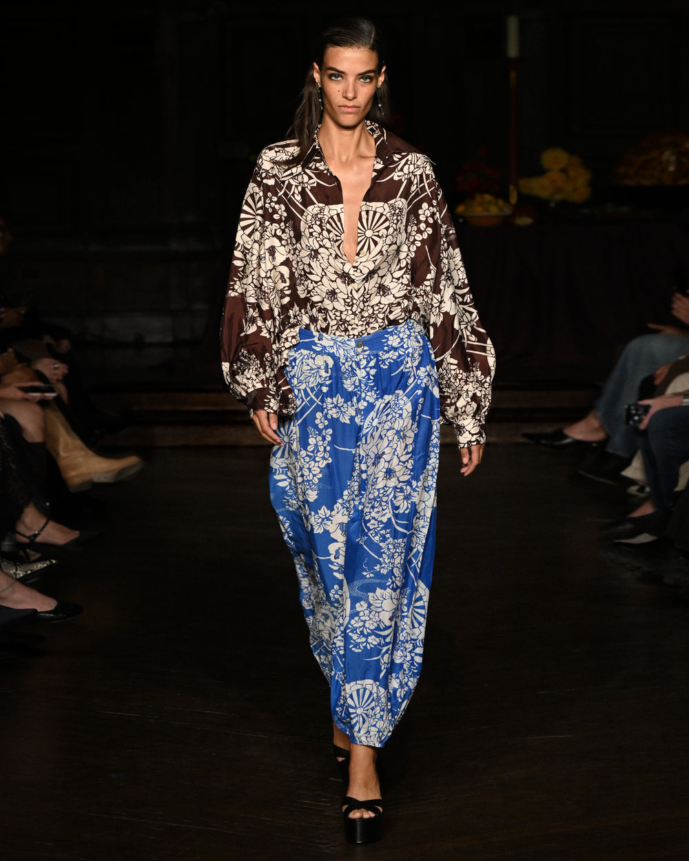 A model walks the runway in the Emery Auburn Spoke Silk Shirt with a plunging neckline and loose blue floral pants, as the audience watches from both sides.