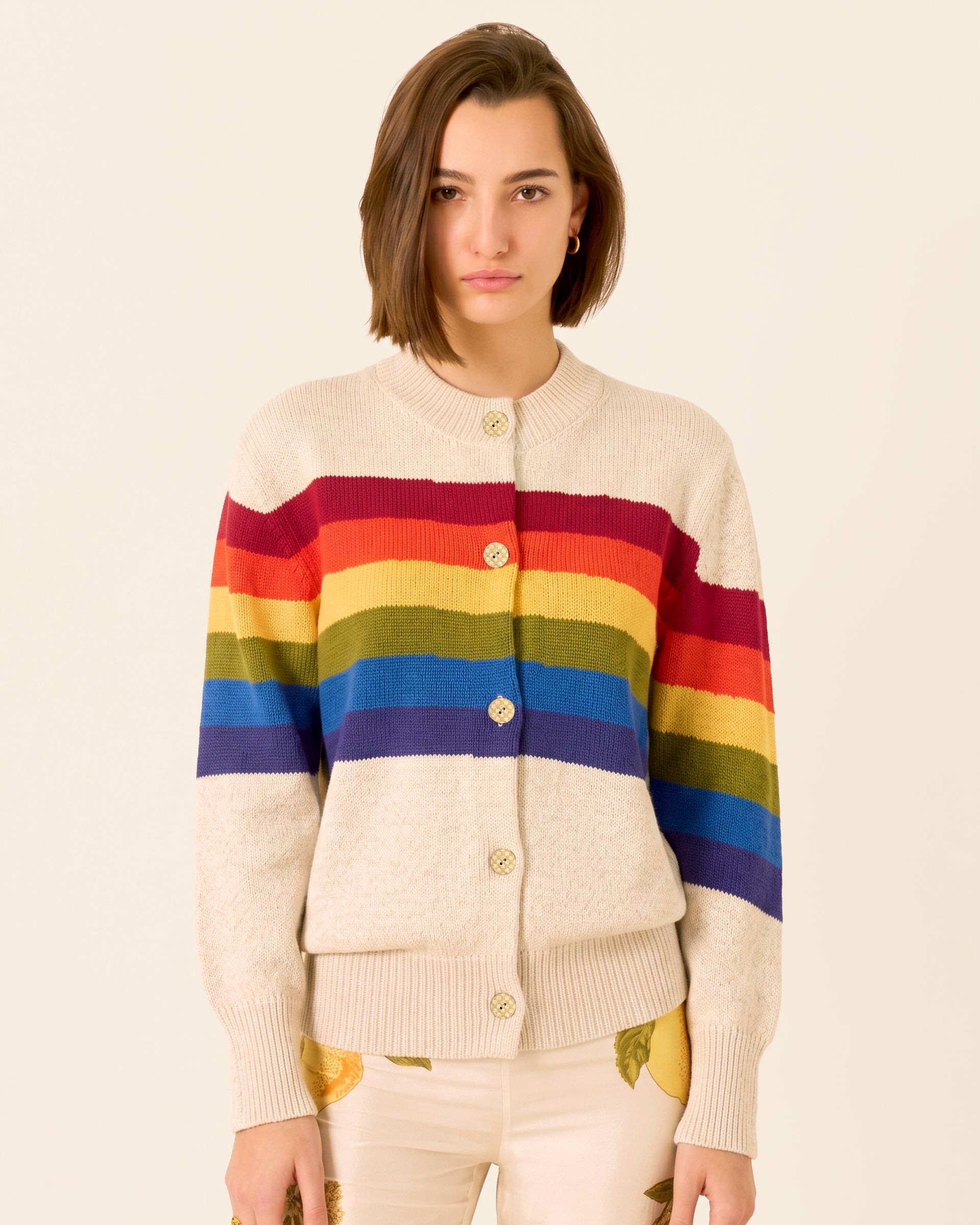 Person wearing the Sammi Prism Knit Cardigan, crafted from cream-colored organic Italian cotton with a rainbow motif, paired with beige pants featuring yellow leaf prints, standing against a plain light background.