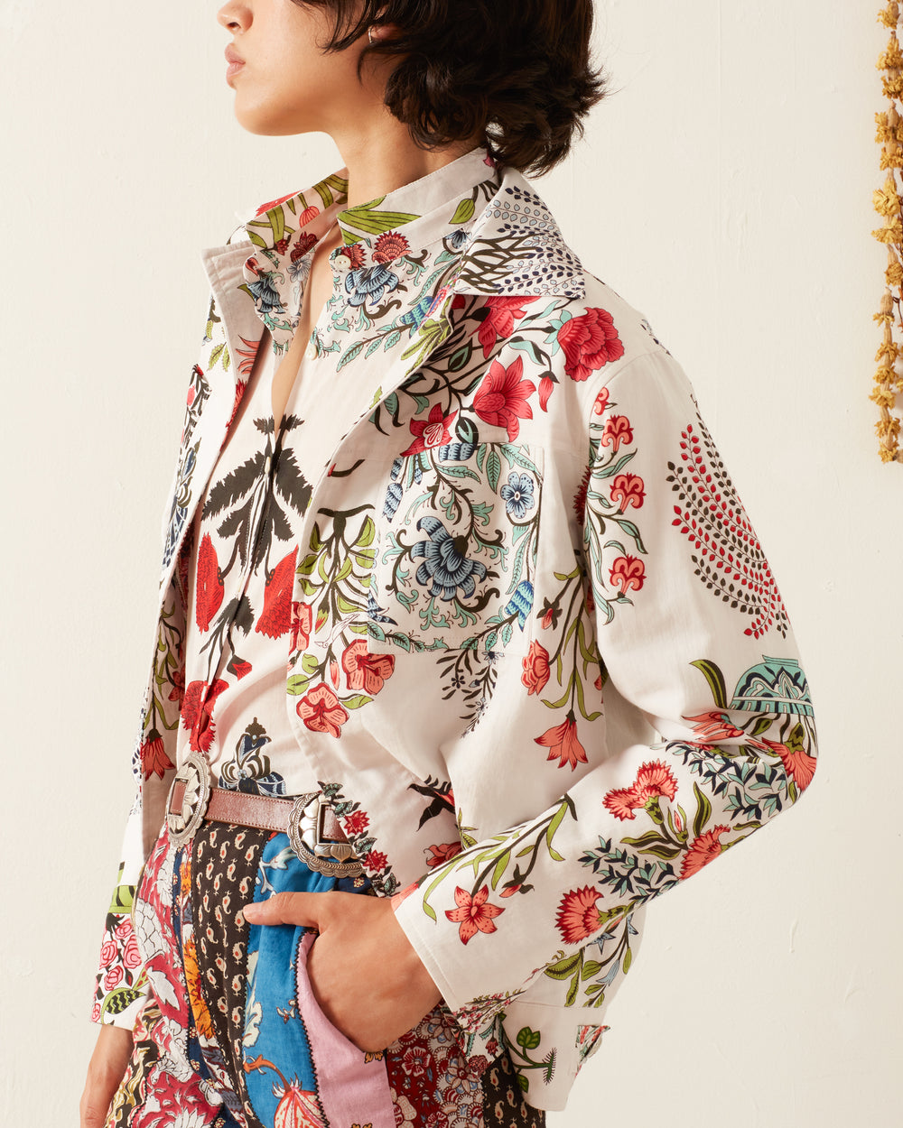 A person dressed in the Seba Flower Show Jacket, known for its colorful hand-printed designs and relaxed fit, paired with a matching dress that highlights intricate patterns. The ensemble features traditional printing techniques in vibrant reds, blues, greens, and other hues, creating a visually striking contrast against the plain background.