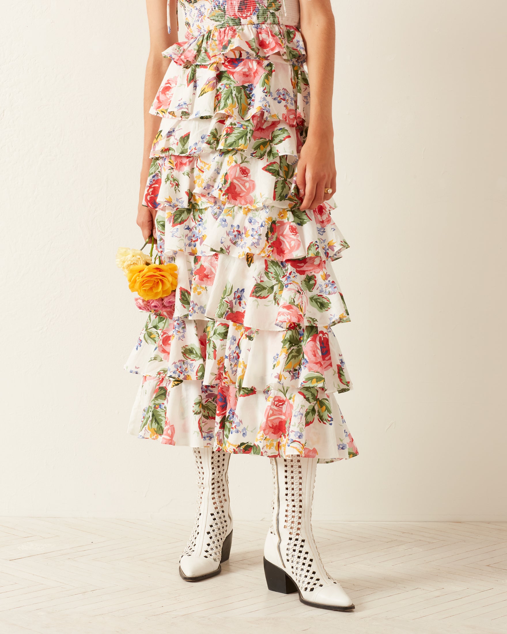 A person dressed in the Tasha Rose Ruffle Skirt embraces a bouquet of yellow flowers. They pair the skirt with white heeled boots featuring cut-out details. The backdrop is a simple, light-colored wall and floor, enhancing the serene ambiance and elegance of their outfit.