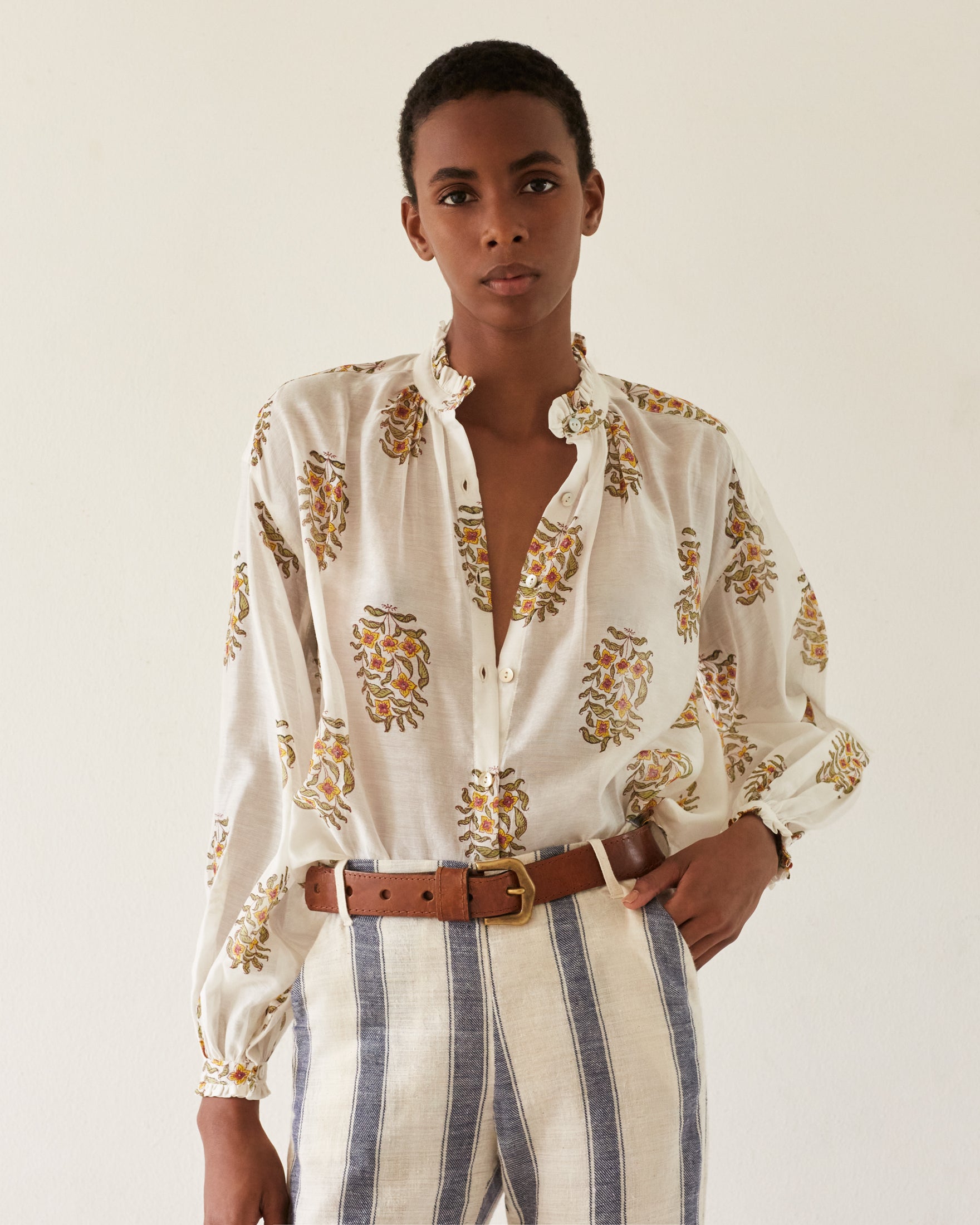 A person wearing a Poet Buttercup Blouse made from hand-printed silk cotton, featuring floral patterns that complement striped pants, stands against a plain background. They have short hair and a brown belt, posing with one hand on their hip.