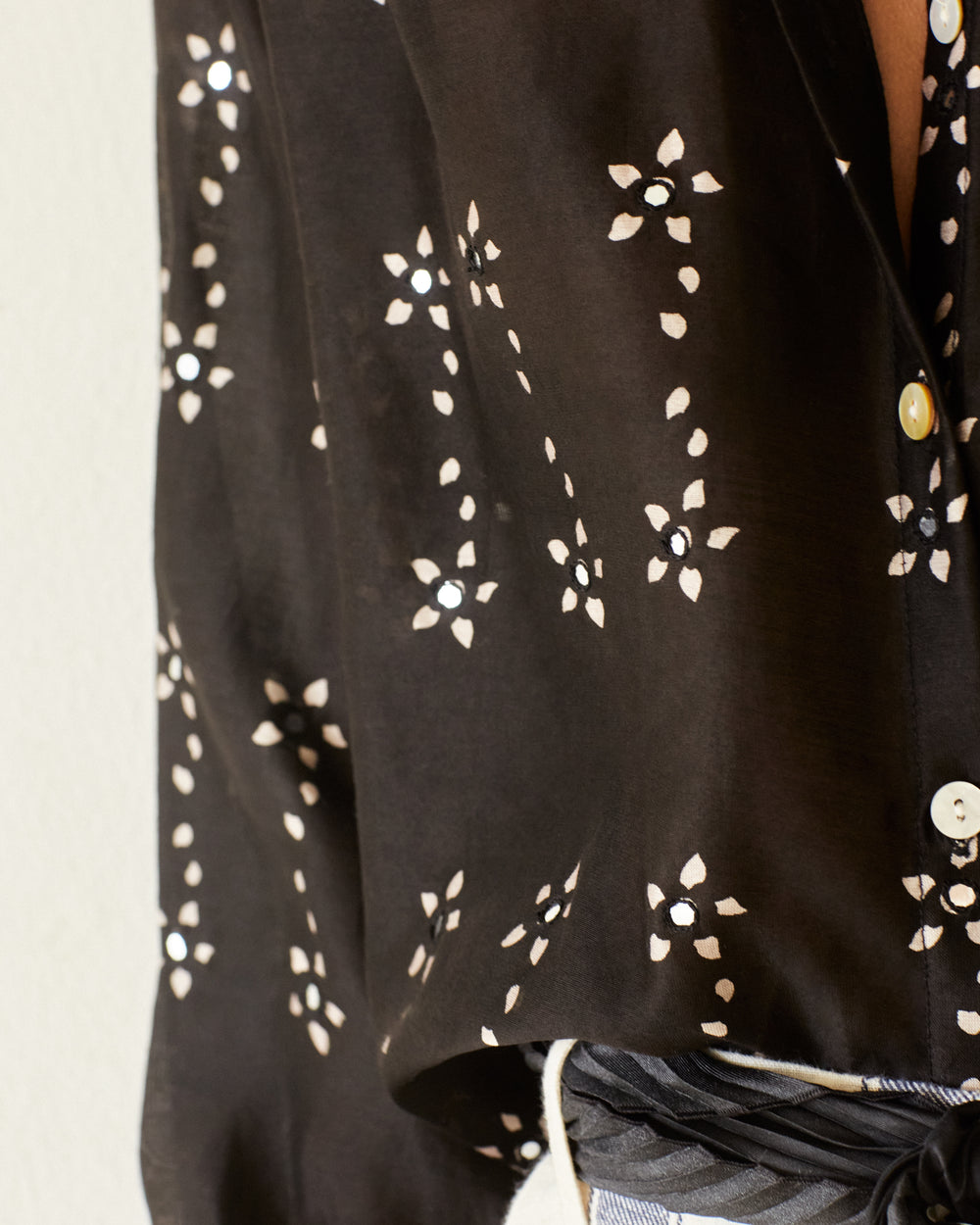 Close-up of a person wearing the Poet Black Daisy Blouse, characterized by its white abstract floral pattern on black, crafted from exquisite silk cotton. The blouse features buttons, with a glimpse of the person's jeans and belt visible at the bottom.