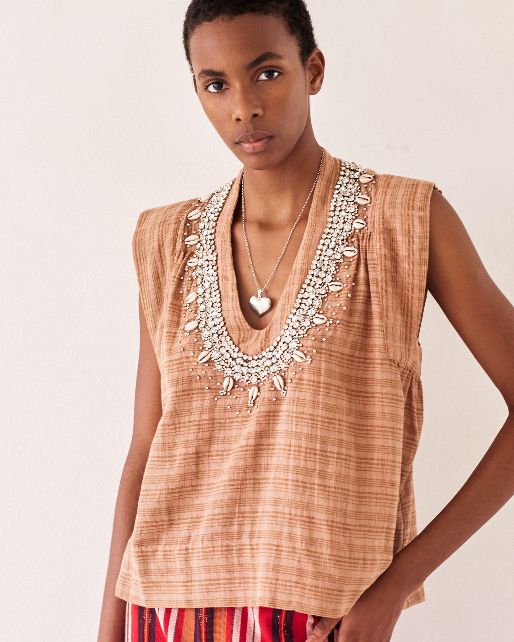 The individual is wearing a light brown Nehru Shell and Gem Top with intricate beadwork around the neckline, complemented by a heart-shaped pendant necklace. This vintage-inspired outfit is paired with a colorful striped skirt, set against a plain, light-colored wall backdrop.