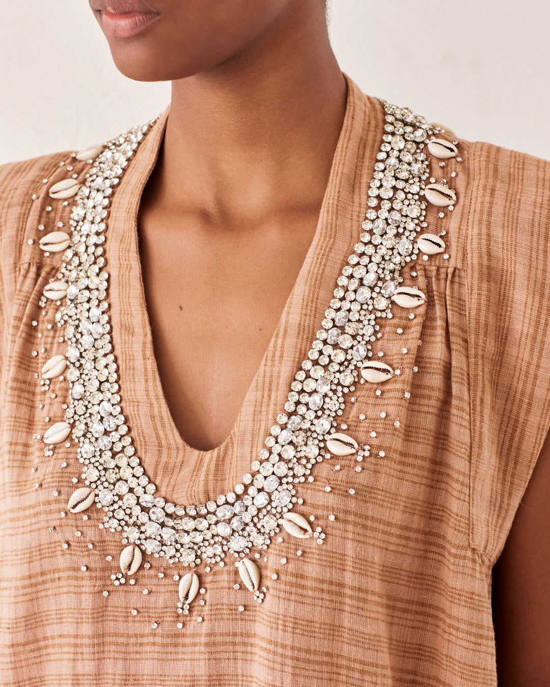 A person wearing the Nehru Shell and Gem Top, a sleeveless brown Bohemian piece featuring a deep V-neck with intricate embellishments of beads, shells, and rhinestones arranged in a striking necklace-like pattern that evokes vintage elegance.