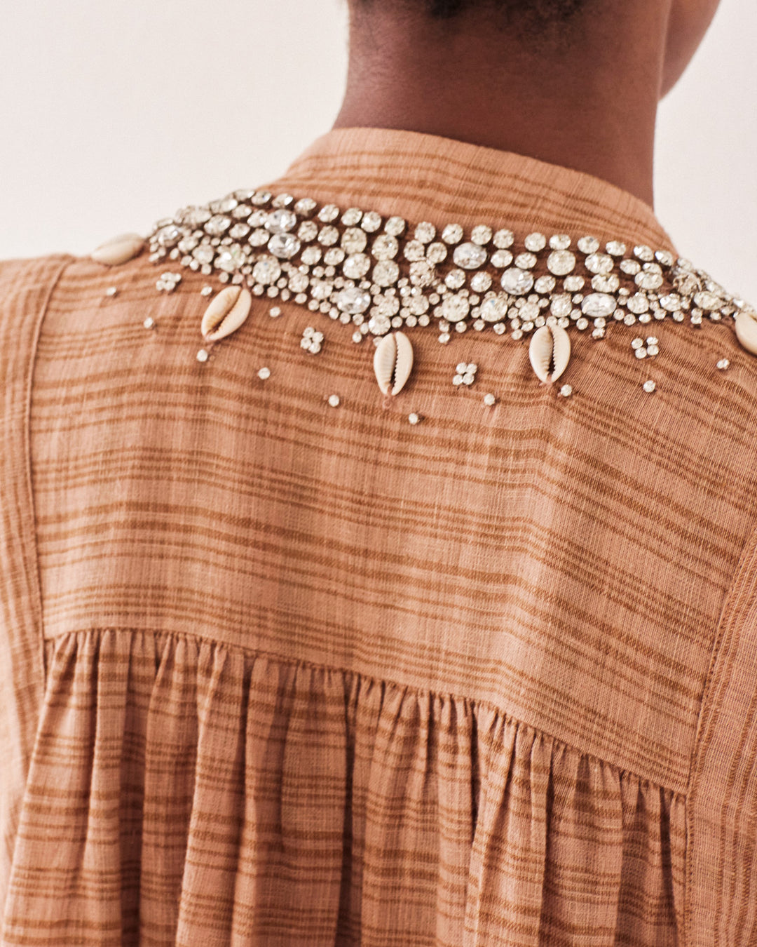 Close-up of a person wearing the Nehru Shell and Gem Top, a textured brown garment adorned with intricate embellishments on the back. This vintage-inspired design features pearls, rhinestones, and shells that form a decorative pattern over the shoulders reminiscent of a bohemian top.