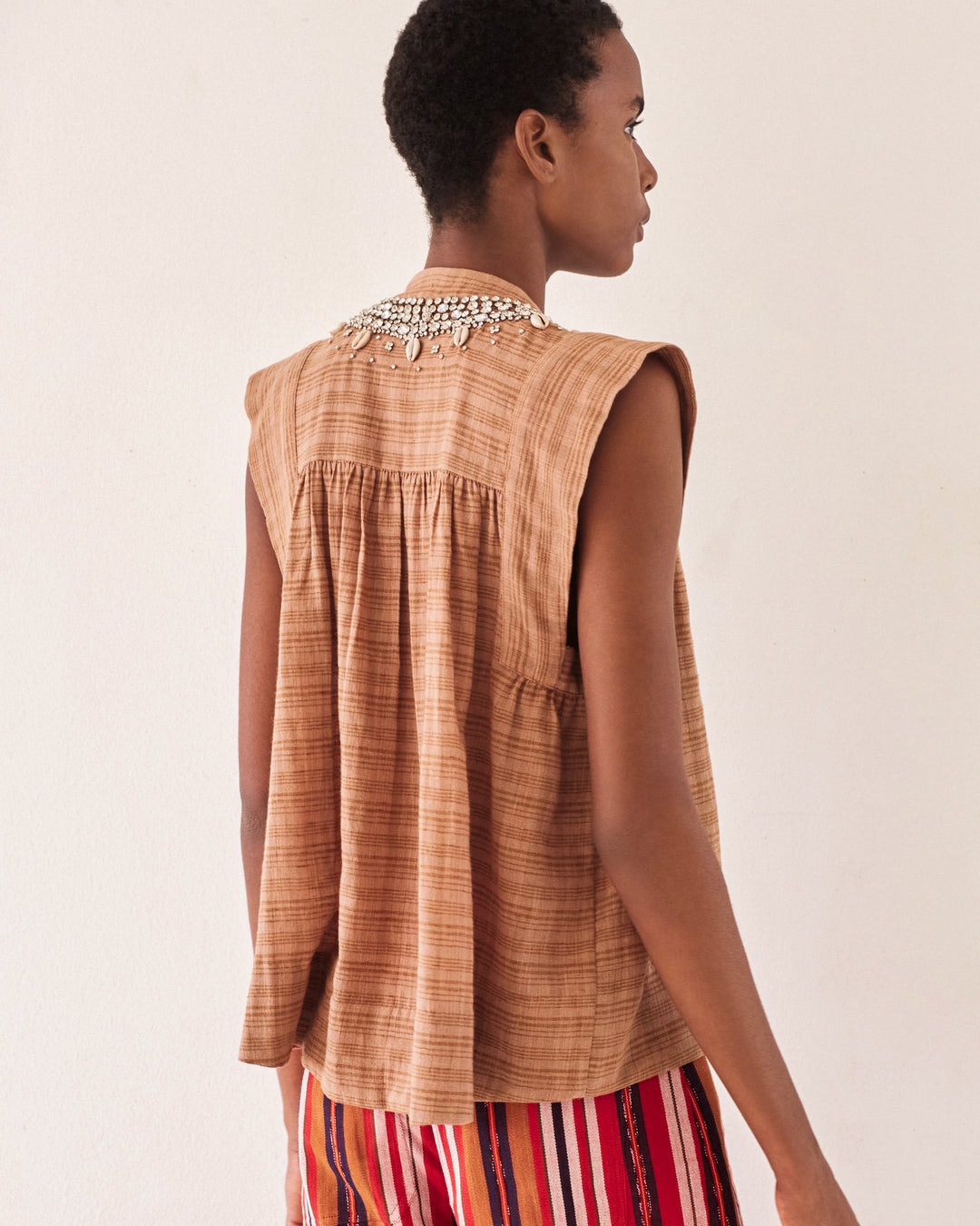 A person wearing the Nehru Shell and Gem Top, a vintage-inspired sleeveless brown piece featuring pleats on the back and adorned with a decorative necklace, stands against a plain white background. Their short hair complements an effortlessly chic Bohemian style vibe.