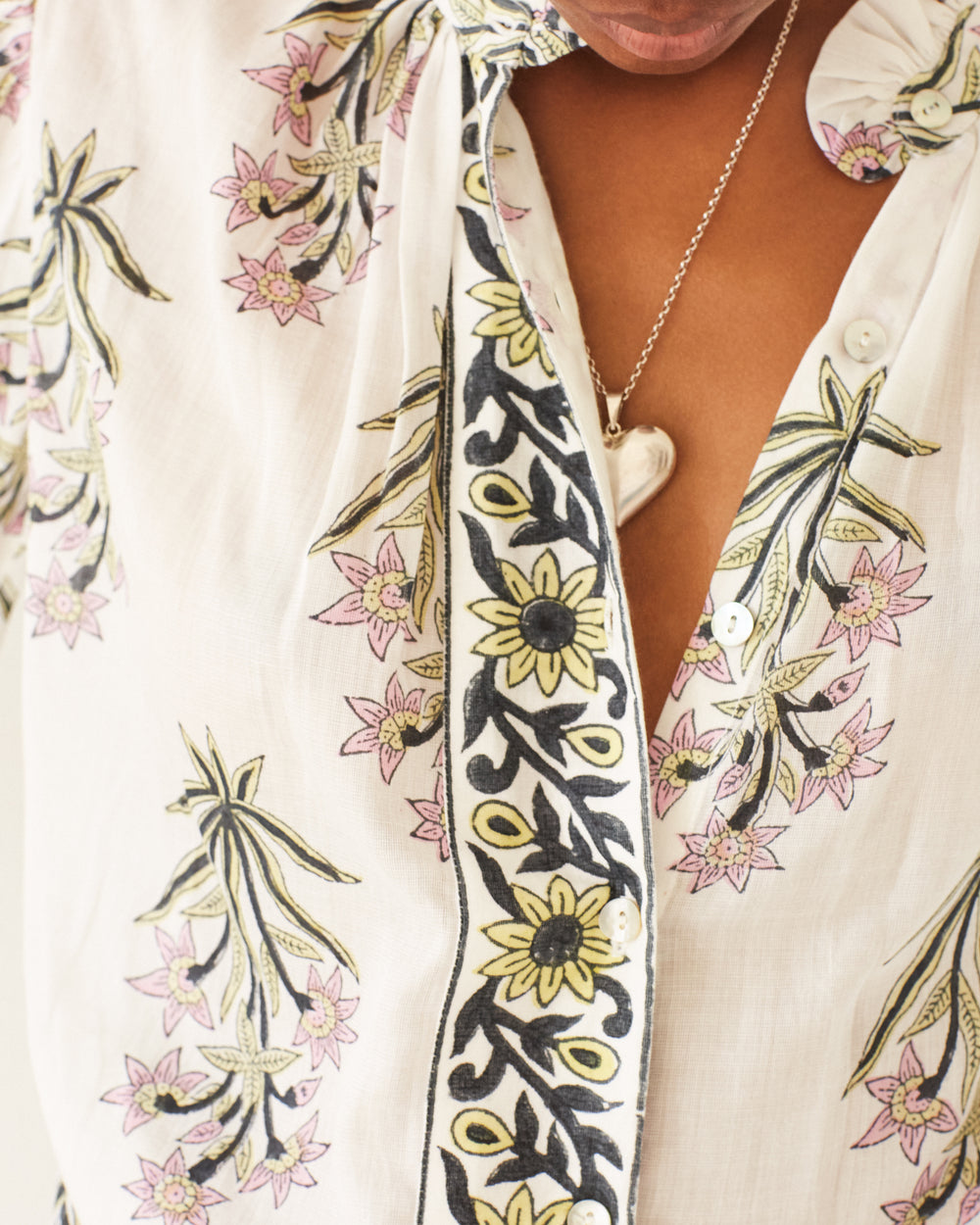 Close-up of a person in an Annabel Winter Lily Shirt, showcasing white floral-patterned fabric adorned with pink and yellow flowers and green leaves. The shirt is partially unbuttoned, highlighting Mother of Pearl buttons and a silver necklace with a heart-shaped pendant. The background is neutral and blurred.