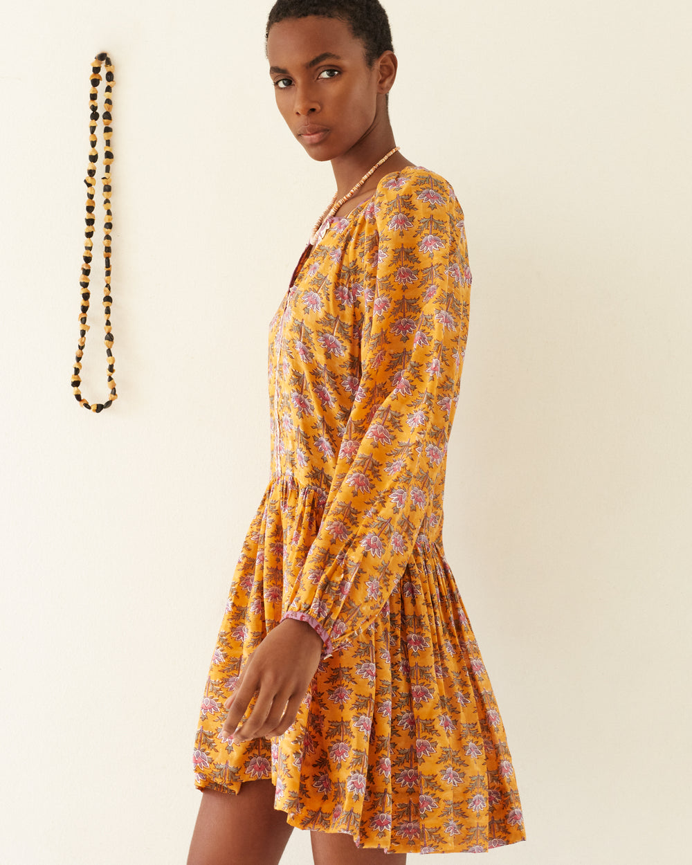 A person with short hair, wearing the Sula Turmeric Dress, stands against a plain wall, looking at the camera. A beaded necklace with mother of pearl accents hangs to their left, adding a touch of elegance to the scene.