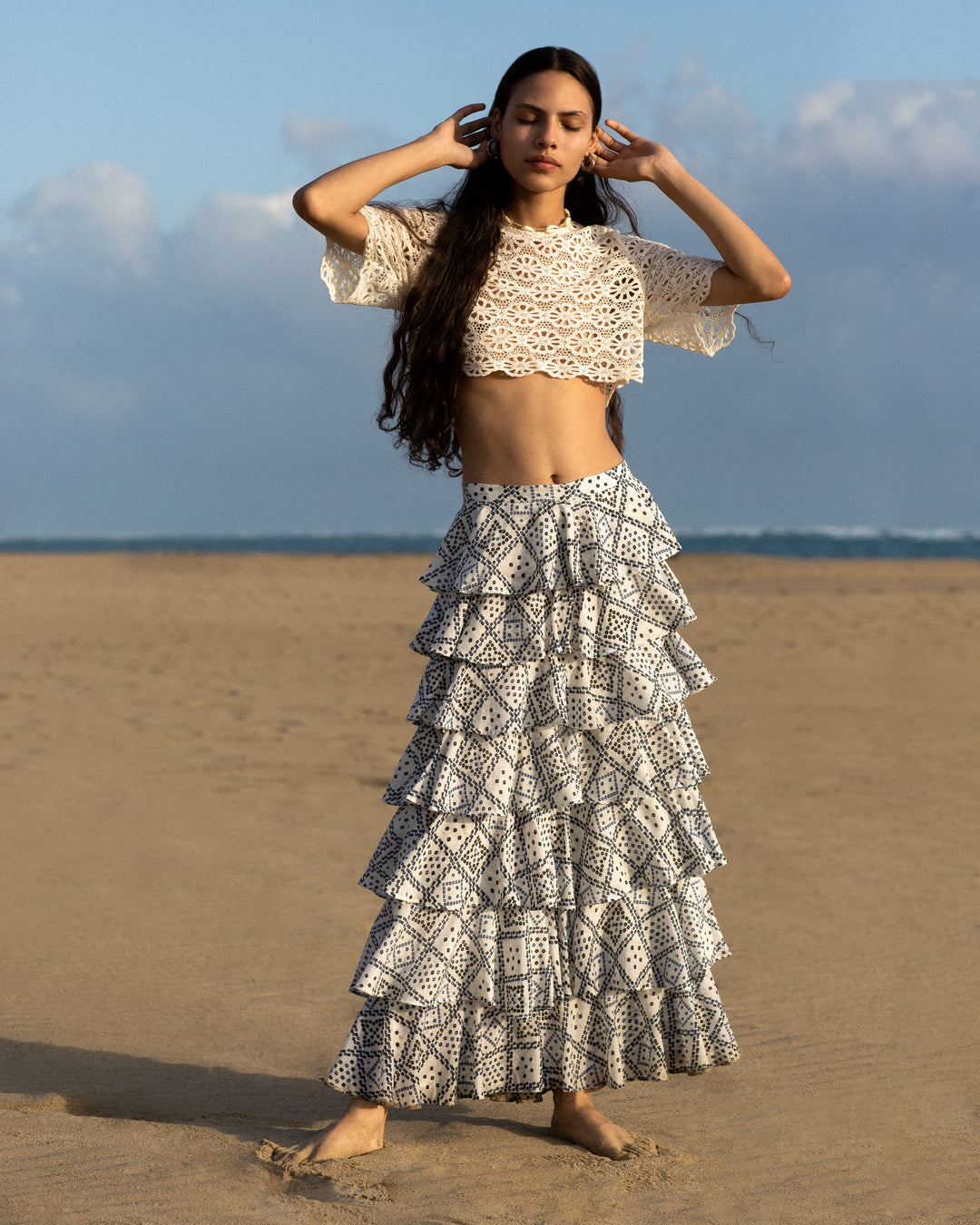 On a sandy beach beneath a clear blue sky, a person with long hair stands, embracing natural beauty in their ensemble. They are wearing a white lace crop top paired with the Tasha Blue Jamdani Skirt, featuring cascading ruffles. With hands near their head, they exude relaxation.